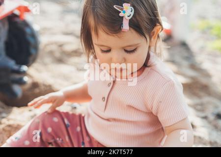 Portrait d'une petite fille assise pensivement dans le jardin et regardant vers le bas concentré sur son jeu. Petit enfant en été. Bonne enfance et Banque D'Images