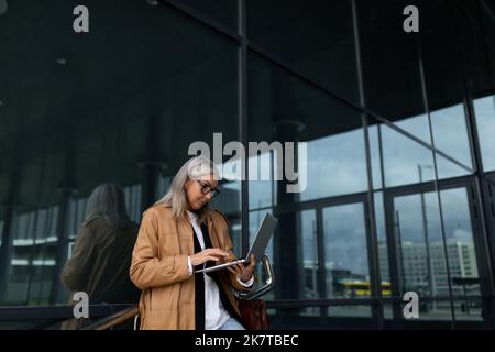 femme élégante de cinquante ans travaillant sur un ordinateur portable à l'entrée du bâtiment de bureau Banque D'Images