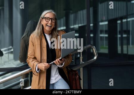 Femme d'affaires de cinquante ans avec un ordinateur portable dans les mains sourit largement en dehors du bureau Banque D'Images