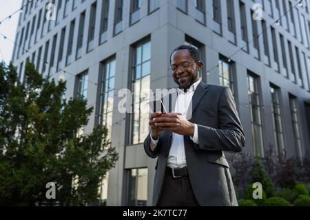 Patron américain africain mature utilisant le téléphone, homme d'affaires à l'extérieur du bâtiment de bureau en costume d'affaires et des lunettes, homme senior tapant le message et la navigation en ligne pages souriant. Banque D'Images