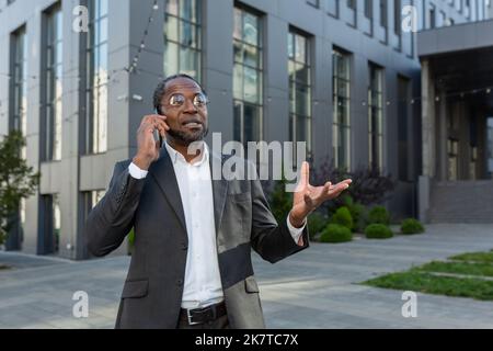 Homme d'affaires senior prospère parlant sur téléphone mobile souriant, patron afro-américain en costume d'affaires et des lunettes sonnant bâtiment de bureau moderne, investisseur parlant à des clients. Banque D'Images