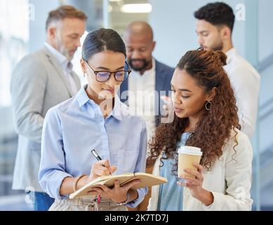 Tout est fait pour faire le travail. deux femmes d'affaires qui passent des notes dans un bureau avec leurs collègues en arrière-plan. Banque D'Images