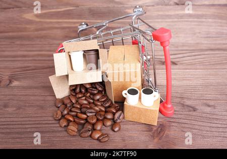 Lot de petites boîtes d'expédition avec des grains de café torréfiés et des tasses par un petit chariot à provisions renversé isolé sur un fond en bois. Banque D'Images