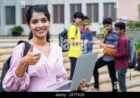 confignce indienne jeune étudiante montrant le pouce vers le haut en regardant la caméra sur le campus d'université tout en utilisant le pc portable - concept de promotion de l'éducation, réussi Banque D'Images