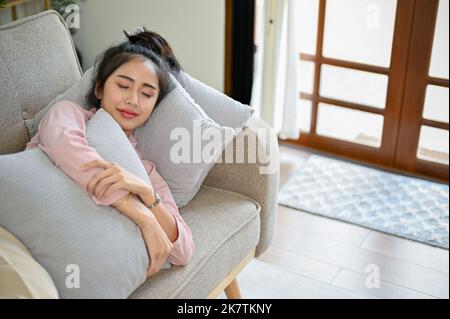 Belle jeune femme asiatique en vêtements décontractés dormant ou prenant la sieste sur son canapé confortable dans son salon. Banque D'Images