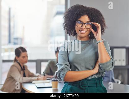 Je ne m'arrête pas. Portrait d'une jeune femme d'affaires repliant ses bras et souriant pendant que ses collègues ont une réunion en arrière-plan. Banque D'Images