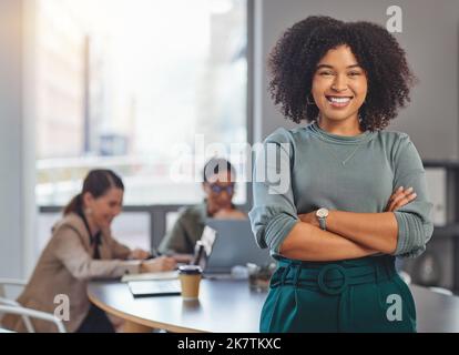Ninja à votre obstacle. Portrait d'une jeune femme d'affaires repliant ses bras et souriant pendant que ses collègues ont une réunion en arrière-plan. Banque D'Images