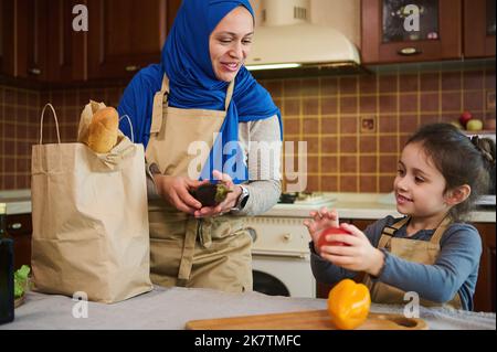 Petite fille aide sa mère à déballer les légumes livrés, prend le poivron, paprika propre frais du sac de papier. Banque D'Images