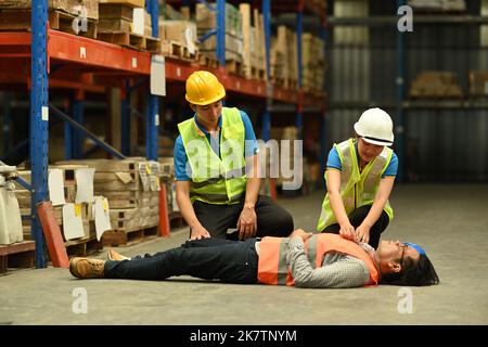 Ouvrier d'entrepôt d'âge moyen allongé inconscient sur le sol en béton et des collègues aidant et donnant les premiers soins blessés Banque D'Images