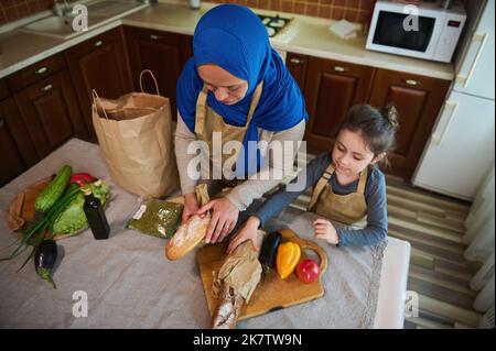 Vue de dessus. Femme musulmane, maman aimante avec la petite fille qui déballe la nourriture du sac de supermarché ensemble pour préparer le repas Banque D'Images