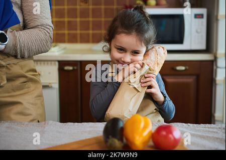 Adorable petite fille tient une baguette chaude de pain de levain de grain entier fraîchement cuit, dans la cuisine avec un design rustique Banque D'Images