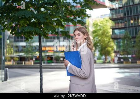 Portrait d'une femme d'entreprise répondant à un appel téléphonique tout en se déplaçant dans le bâtiment du bureau. Femme d'affaires qui appelle quelqu'un, tient des documents Banque D'Images