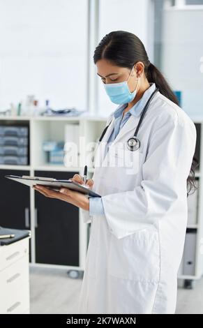 Remplir une carte covid du patient. un médecin portant un masque facial et debout seul en écrivant sur son presse-papiers dans sa clinique. Banque D'Images