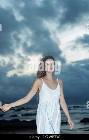 Belle femme vêtue d'une robe blanche marchant sur la plage un jour d'été nuageux Banque D'Images