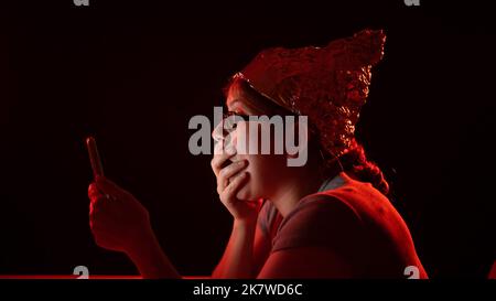 Femme caucasienne paranoïaque utilisant un smartphone portant un chapeau Tinfoil. Théorie du complot. Banque D'Images