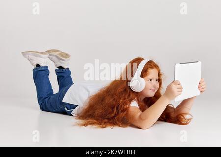 Regarder des dessins animés. Jolie petite fille à tête rouge portant un tee-shirt et un Jean avec une tablette isolée sur fond blanc de studio. Émotions des enfants, visage Banque D'Images