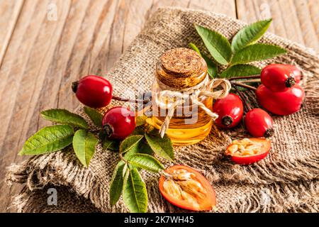 une bouteille en verre avec un liège avec de l'huile de graines de rosehip naturelle sur un terlap et une table de village parmi les hanches de roses fraîchement cueillies. produit biologique Banque D'Images