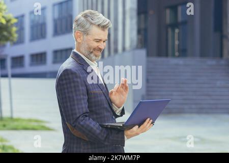 Un patron d'âge mûr parlant debout lors d'un appel vidéo, un homme d'affaires aux cheveux gris tenant un netbook regardant une webcam et expliquant comment discuter, un homme en voyage d'affaires suit un immeuble de bureau avec un ordinateur portable. Banque D'Images