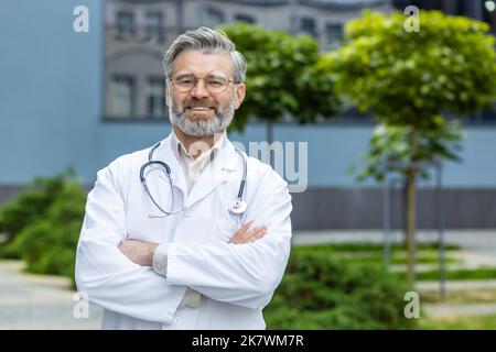 Portrait d'un médecin âgé aux cheveux gris, d'un homme mature en manteau médical blanc avec bras croisés et stéthoscope souriant et regardant la caméra, médecin-chef à l'extérieur de l'hôpital moderne. Banque D'Images