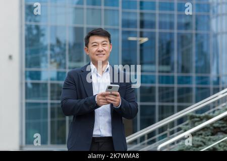 Portrait d'un homme d'affaires asiatique à l'extérieur d'un immeuble de bureaux moderne, patron en costume d'affaires en utilisant le téléphone souriant et regardant l'appareil photo, homme tapant des messages et de parcourir des pages Web en ligne. Banque D'Images