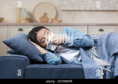 Maladie. Quarantaine. Une jeune belle femme asiatique est couché à la maison sur le canapé, couvert d'une couverture, malade. Il a un nez qui coule, essuie son nez avec une serviette. Il se sent mal. Banque D'Images