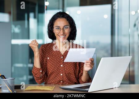 Portrait d'une femme heureuse, une femme hispanique a reçu une lettre de message heureuse et regardant l'appareil photo, la femme d'affaires en lunettes et les cheveux bouclés travaillant à l'intérieur d'un immeuble de bureau moderne à l'aide d'un ordinateur portable. Banque D'Images