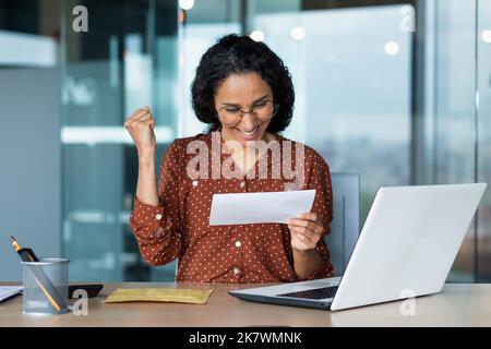 Une femme hispanique a reçu une lettre heureuse de la banque, une femme d'affaires lit et se réjouit de célébrer le succès, une femme travaillant dans des lunettes et des cheveux bouclés dans un immeuble de bureau moderne utilise un ordinateur portable. Banque D'Images