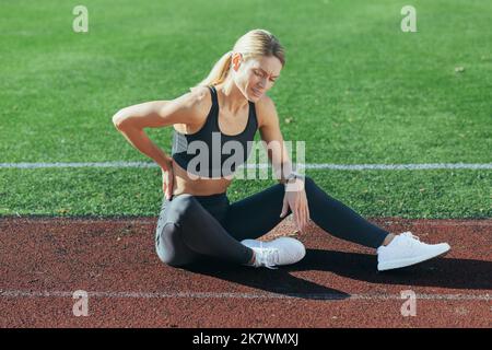 Une athlète féminine blessée est assise sur le tapis roulant du stade, a de graves douleurs au dos, tient sa main à ses côtés, masse un muscle. Banque D'Images
