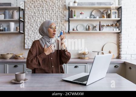 Femme asthmatique à la maison, femme musulmane en hijab utilisant un inhalateur pour soulager la respiration, femme au foyer travaillant à distance à la maison en utilisant un ordinateur portable, ayant de la difficulté à respirer à la maison. Banque D'Images