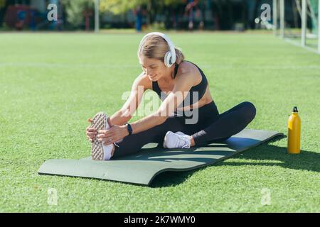 Femme blonde faisant des exercices physiques assis sur un tapis de sport, sportswoman étirant les muscles, écoutant de la musique dans les écouteurs et souriant, stade le matin par beau temps. Banque D'Images