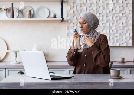 Femme asthmatique à la maison, femme musulmane en hijab utilisant un inhalateur pour soulager la respiration, femme au foyer travaillant à distance à la maison en utilisant un ordinateur portable, ayant de la difficulté à respirer à la maison. Banque D'Images