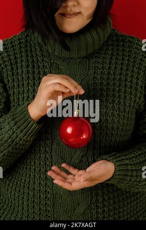 Femme asiatique dans un chandail vert sur fond rouge tient une boule de Noël rouge dans ses mains. Pas de face. Le concept de la préparation de Noël et de la décoration Banque D'Images