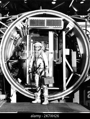 L'astronaute John H. Glenn Jr., adapté avec un tuyau pour s'adapter à l'unité de ventilation fixée, pendant l'essai en chambre d'altitude. Il est debout à l'entrée de la chambre d'essai avec sa visière de casque vers le bas. Banque D'Images