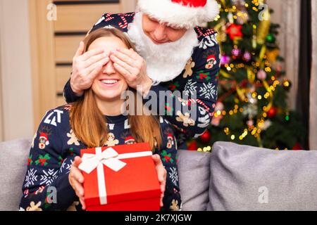 Un homme habillé comme père Noël couvrant les yeux de sa petite amie pendant que lui donne la surprise de Noël. Homme donnant cadeau de noël à sa petite amie Banque D'Images