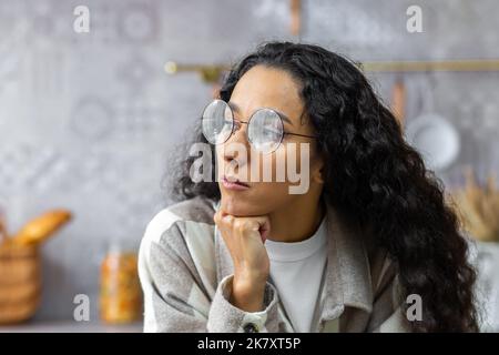 Triste femme hispanique gros plan à la maison, portrait de la femme bouleversée avec des lunettes et des cheveux bouclés regardant par la fenêtre Banque D'Images