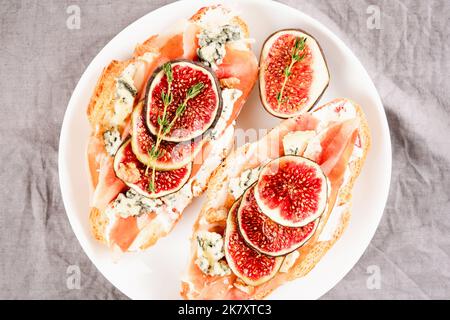 Bruschetta croissant frais avec figue mûre, fromage bleu, prosciutto, noix et herbes sur plaque blanche. Mise au point sélective Banque D'Images