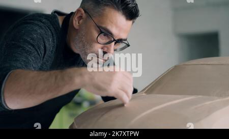 Un concepteur automobile expérimenté fait glisser l'outil de façonnage en bois pour lisser la surface du prototype de sculpture de voiture. Concepteur avec un tablier travaille sur la sculpture en argile de la voiture. Banque D'Images