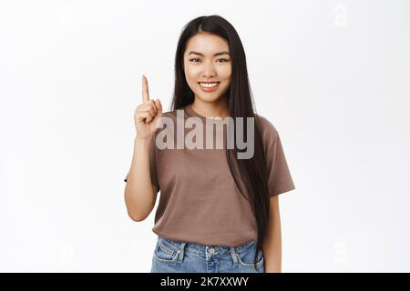 Portrait d'une fille coréenne souriante montrant une offre promotionnelle, une bannière publicitaire, porte un t-shirt, se dresse sur fond blanc Banque D'Images