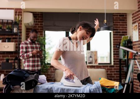 Femme chantant bruyamment avec effusivité tout en faisant des corvées à la maison, le mari afro-américain rit amusé dans l'écoute de fond. Femme au foyer faisant volontiers le ménage, couple culturellement divers. Banque D'Images