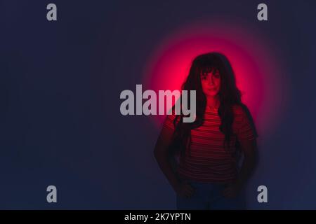 Portrait artistique créatif d'une femme aux cheveux foncés debout devant un mur dans l'obscurité avec un spot rouge sur son visage. Prise de vue horizontale en studio. Photo de haute qualité Banque D'Images