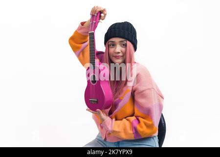 Jeune fille blanche avec des cheveux longs roses portant un chandail coloré assis dans la chaise montrant le ukulele rose à l'appareil-photo. Arrière-plan blanc isolé. Prise de vue horizontale en studio. Photo de haute qualité Banque D'Images