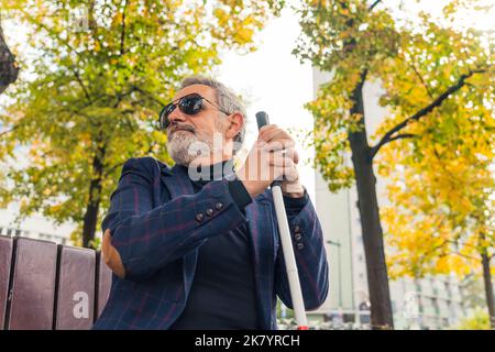 Homme aveugle d'âge moyen avec cheveux gris et barbe dans des lunettes de soleil assis sur un banc dans le parc de la ville tenant la canne blanche regardant autour. Prise de vue horizontale en extérieur. Photo de haute qualité Banque D'Images