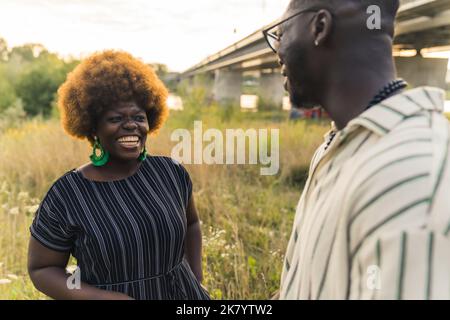 Charmant couple afro-américain debout au bord de la rivière, au milieu de la prairie, en riant et en flirtant les uns avec les autres. Relations humaines. Deux Noirs - une femme et un homme - dans des vêtements dépouillés et décontractés qui parlent. Photo de haute qualité Banque D'Images