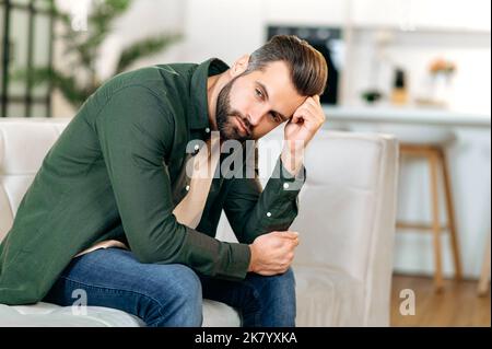 Dépression, fatigue, frustration. Contrarié homme caucasien, asseyez-vous sur un canapé dans la salle de séjour, penché sur la main, éprouvant des problèmes personnels, le stress, la solitude, a besoin de soutien psychologique et de repos Banque D'Images