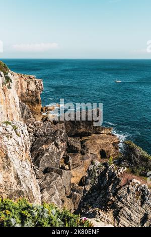 Un paysage vertical d'une falaise rocheuse avec une végétation clairsemée, beaucoup de pierres énormes, et un bateau solitaire derrière le rivage flottant sur les vagues turquoise Banque D'Images