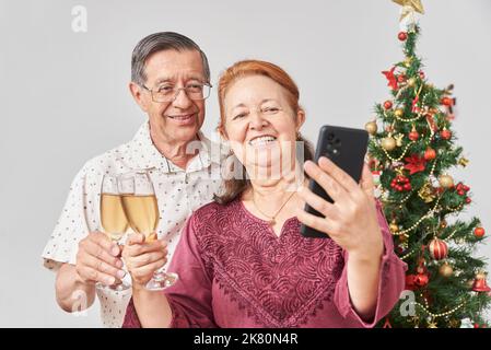 Heureux couple latin senior faisant un selfie avec un smartphone tout en toaster pendant Noël ou le nouvel an, célébrant des vacances à la maison, ayant un bon t Banque D'Images