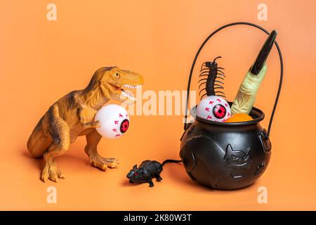 Jouet dinosaure Tyrannosaurus tenant l'œil dans ses pattes, bol avec chauve-souris, citrouille isolée sur fond orange carte de voeux de vacances joyeux jour d'Halloween cre Banque D'Images
