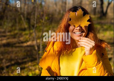 Femme caucasienne aux cheveux rouges tenant une feuille d'érable tombée.Marche d'automne. Banque D'Images