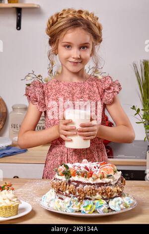 La petite fille fait un gâteau fait maison avec une recette facile à la cuisine contre un mur blanc avec des étagères dessus. Banque D'Images