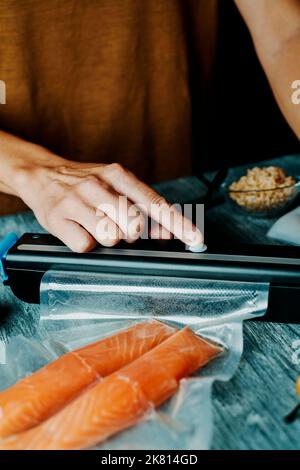 un jeune homme caucasien emboîte sous vide deux morceaux de saumon dans un sac transparent, sur une table rustique grise Banque D'Images